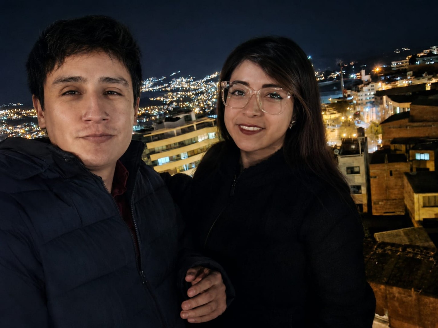 Amy y Ricardo juntos de noche, con las luces de la ciudad al fondo.